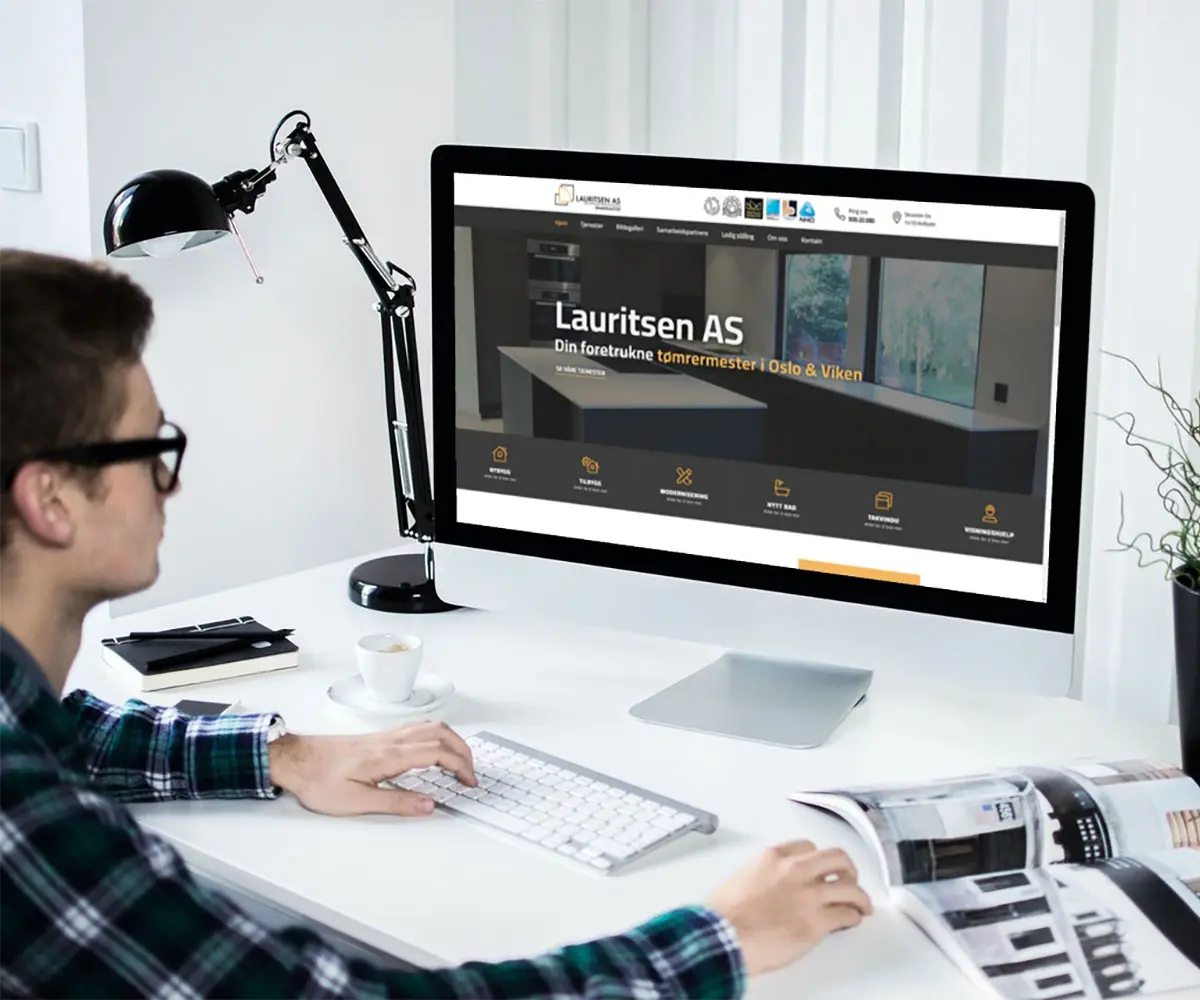 Bilde av en person som jobber på sitt skrivebord med en Mac skjerm. På skjermen er det Lauritsen As sin hjemmeside, i samarbeid med UndrumDesign.