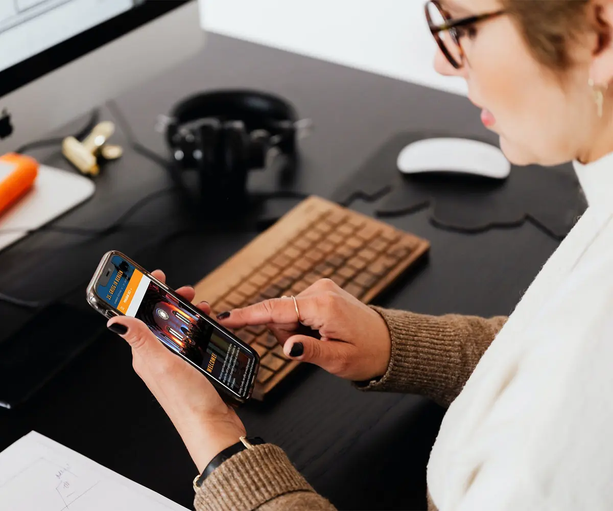 Bilde av en person som ved skrivebordet sitt som holder en mobiltelefon. På skjermen er det GriegInBergen sin hjemmeside, i samarbeid med UndrumDesign.