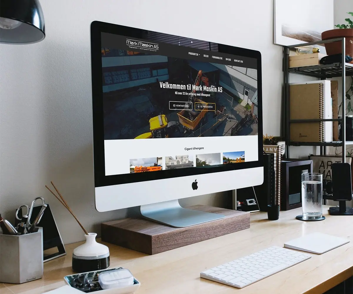 Bilde av en Mac Skjerm på skrivebord med diverse elementer. På skjermen er det Mørk Maskin AS sin hjemmeside, i samarbeid med UndrumDesign.